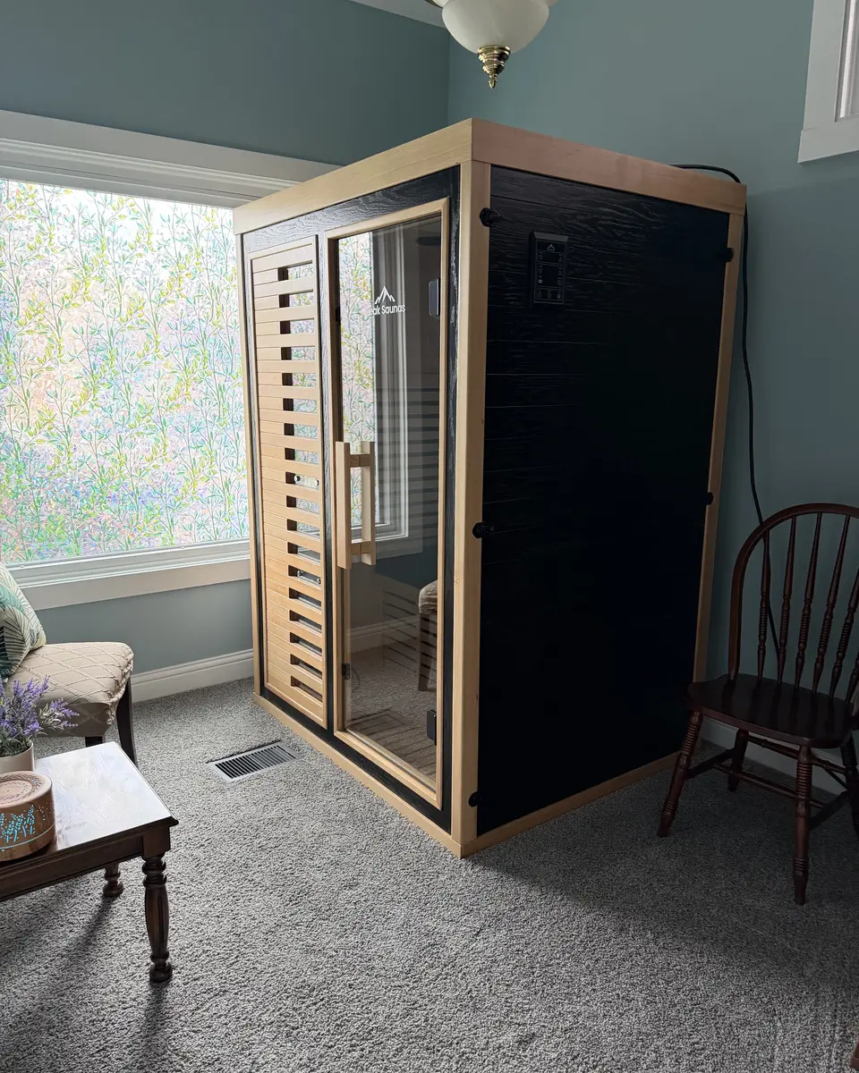 A light-wood sauna cabin with a glass door, a wicker chair beside it, in a quiet blue-gray room.