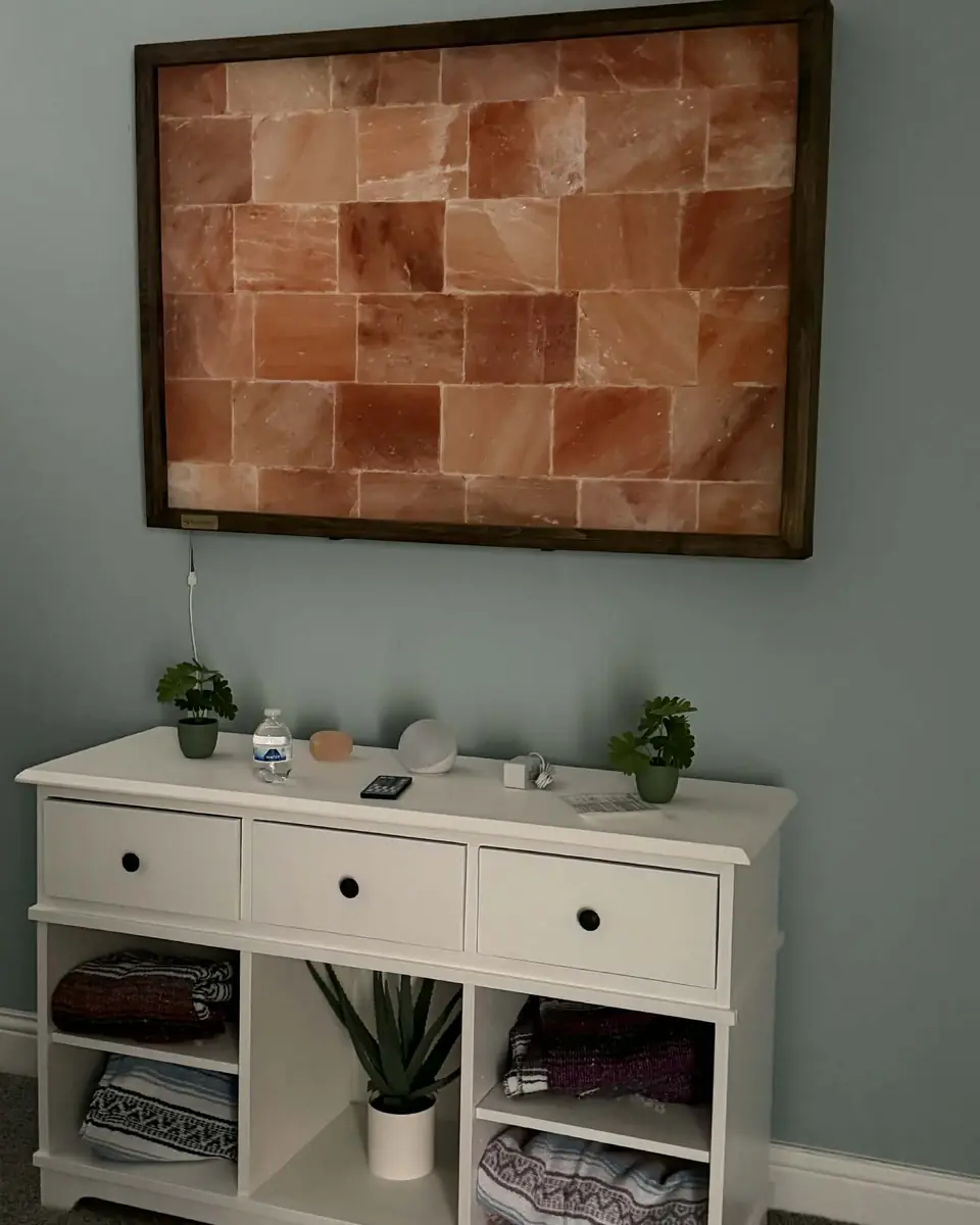 A framed panel of backlit Himalayan salt bricks above a white console holding folded blankets and a small plant.